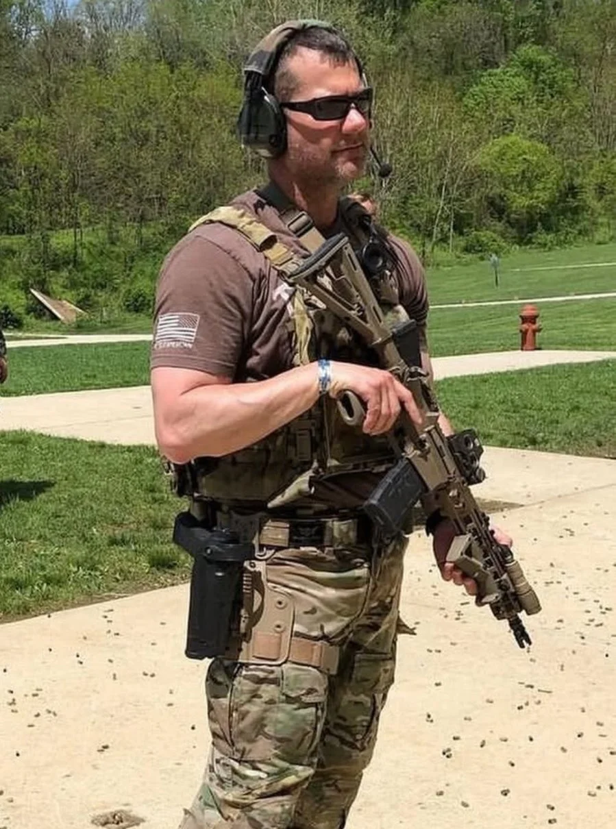A man dressed in military tactical gear, wearing sunglasses and protective ear gear, stands outdoors on a paved surface with a grassy background, holding a rifle.