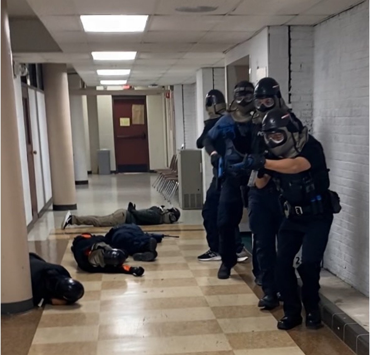 Police officers in riot gear standing in a hallway near fallen backpacks and a person lying on the floor in a school or public building.