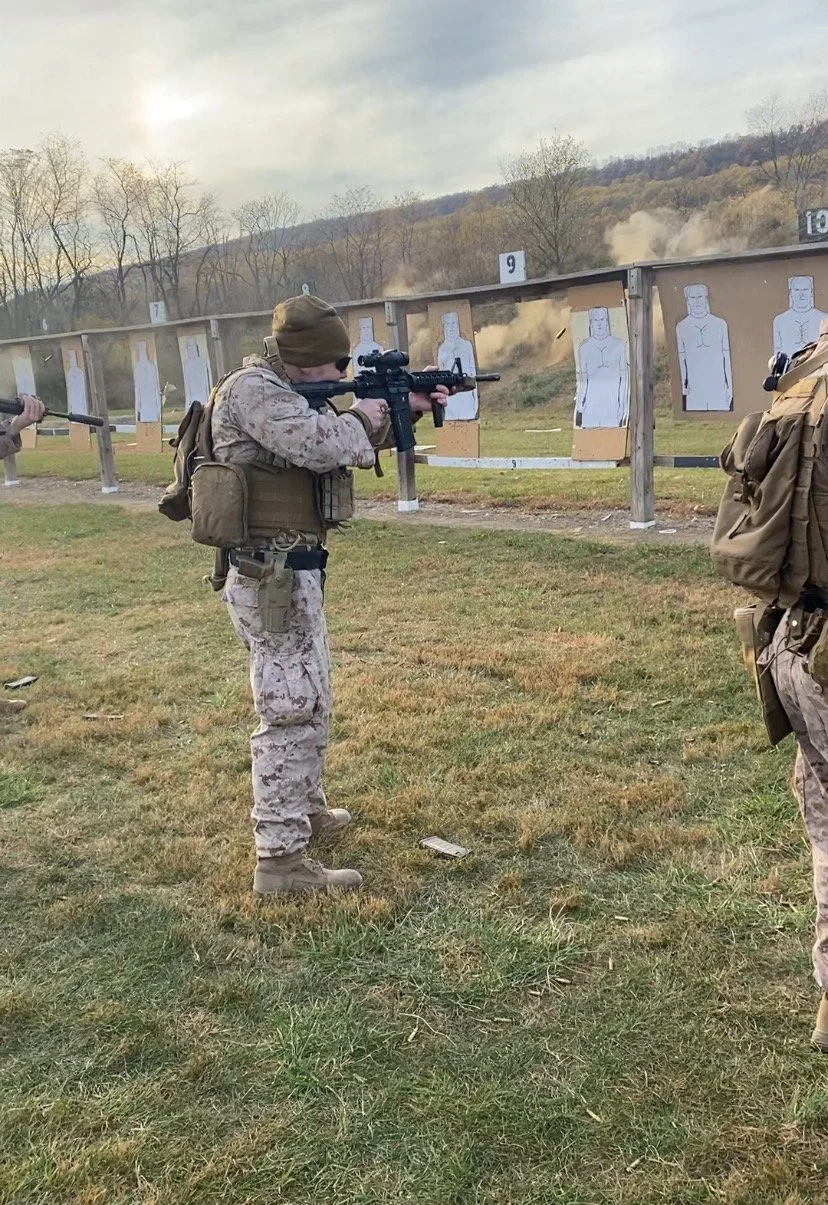 Soldier aiming a rifle at shooting targets with paper silhouettes, outdoor shooting range with hills and trees in the background, cloudy sky.