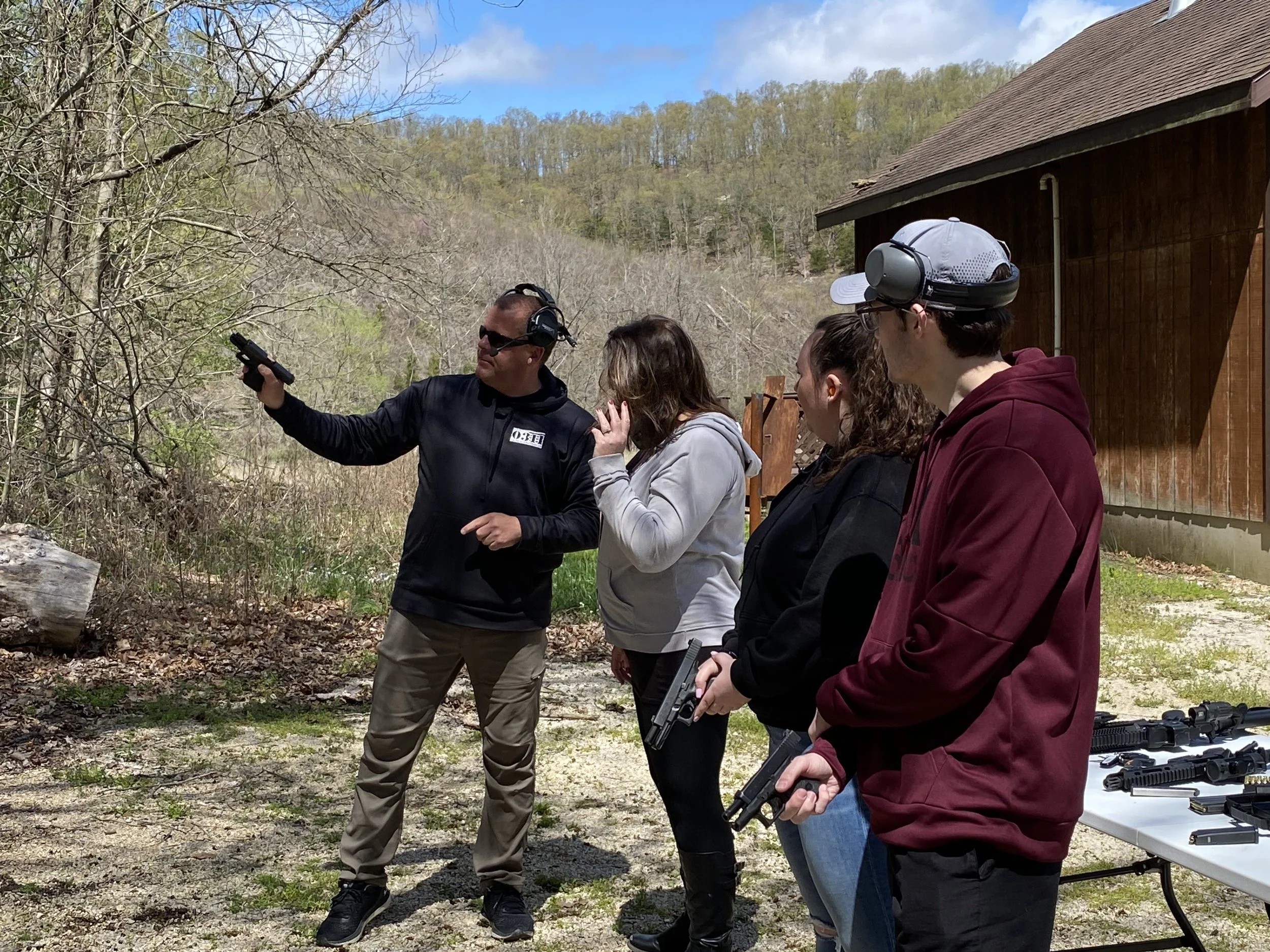Instructor demonstrating firearm safety to students outdoors, with guns and firearm accessories on a table nearby.