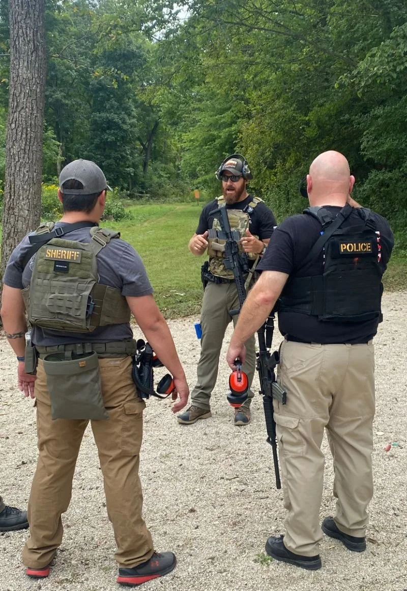 Three law enforcement officers, two with sheriff and one with police, standing outdoors on a gravel path in a wooded area, engaged in conversation. One officer is speaking and holding a rifle, while the others are listening. All officers are wearing 