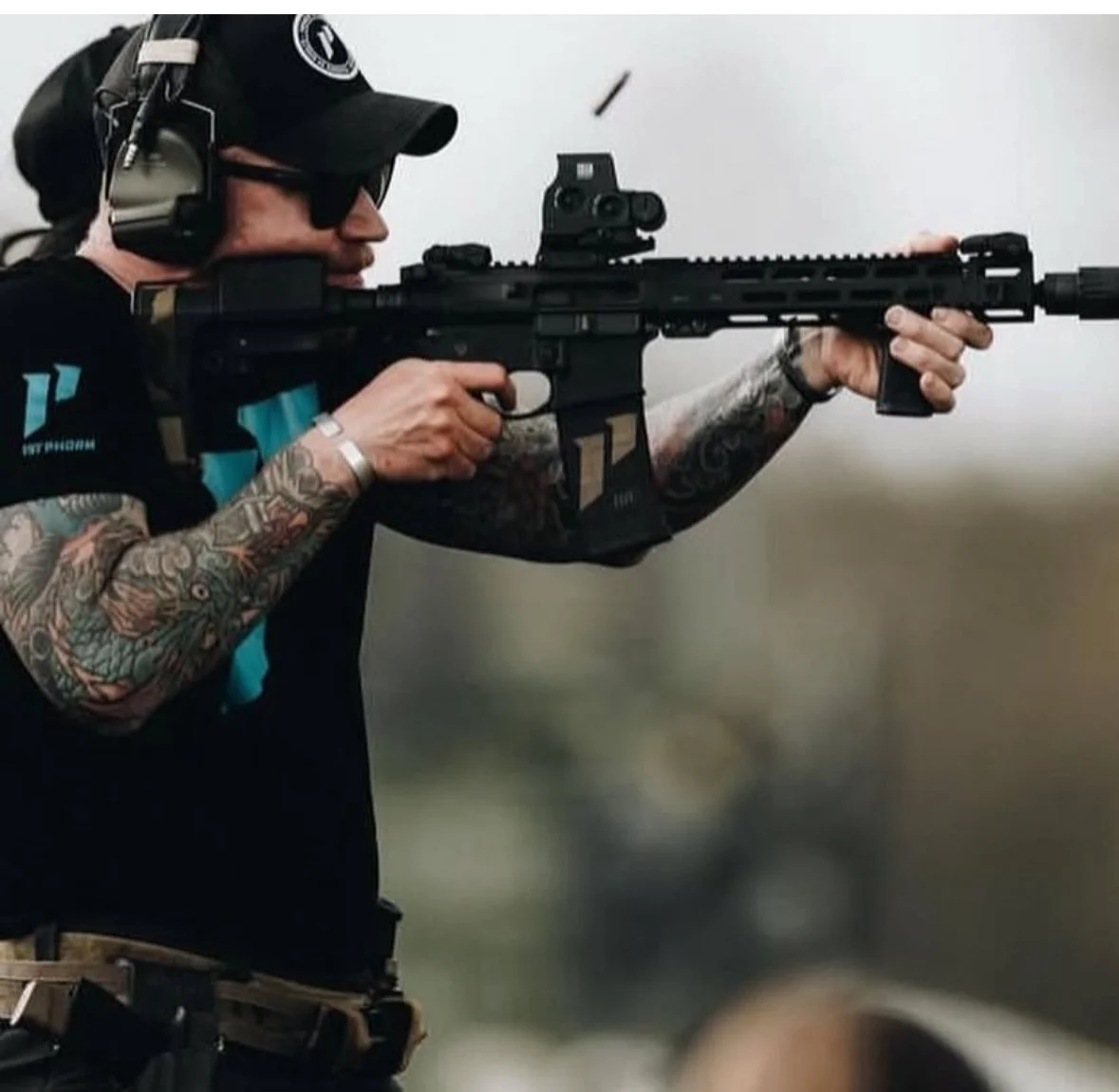 Person with tattoos shooting a rifle with a scope, wearing a cap and headphones, in an outdoor setting.