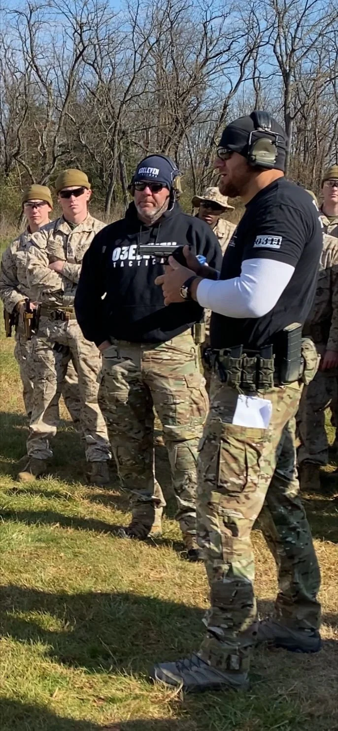 A group of soldiers in camouflage uniforms standing outdoors, listening to a man in tactical gear and a headset explaining something. One soldier is holding a gun, and some are wearing sunglasses and hats.