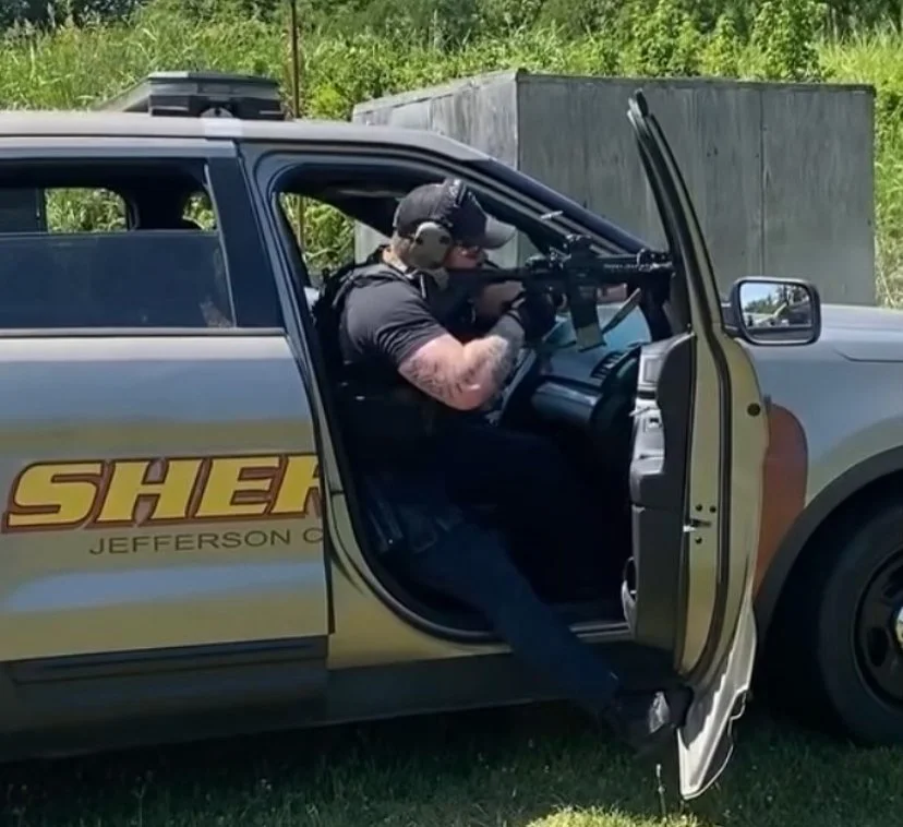 A sheriff deputy in uniform and tactical gear aiming a rifle from the open door of a sheriff patrol car, parked on grass with trees in the background.