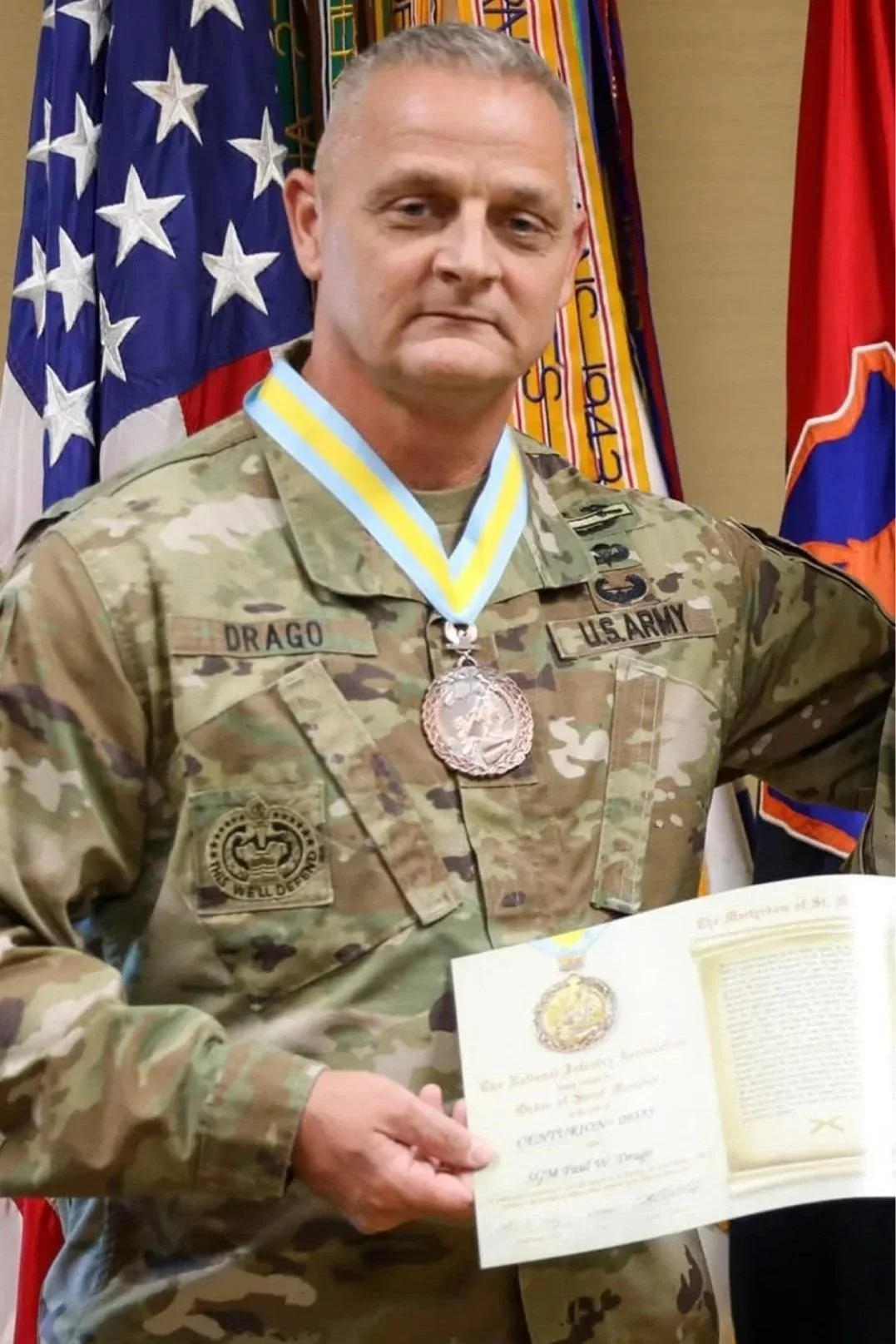 A man in military uniform holding a certificate, with American and military flags in the background.