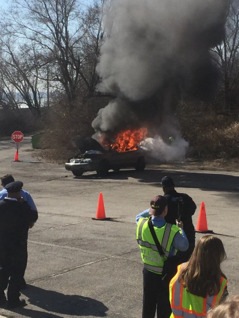 Car on fire with black smoke, surrounded by police and safety personnel, orange traffic cones, and a stop sign nearby.