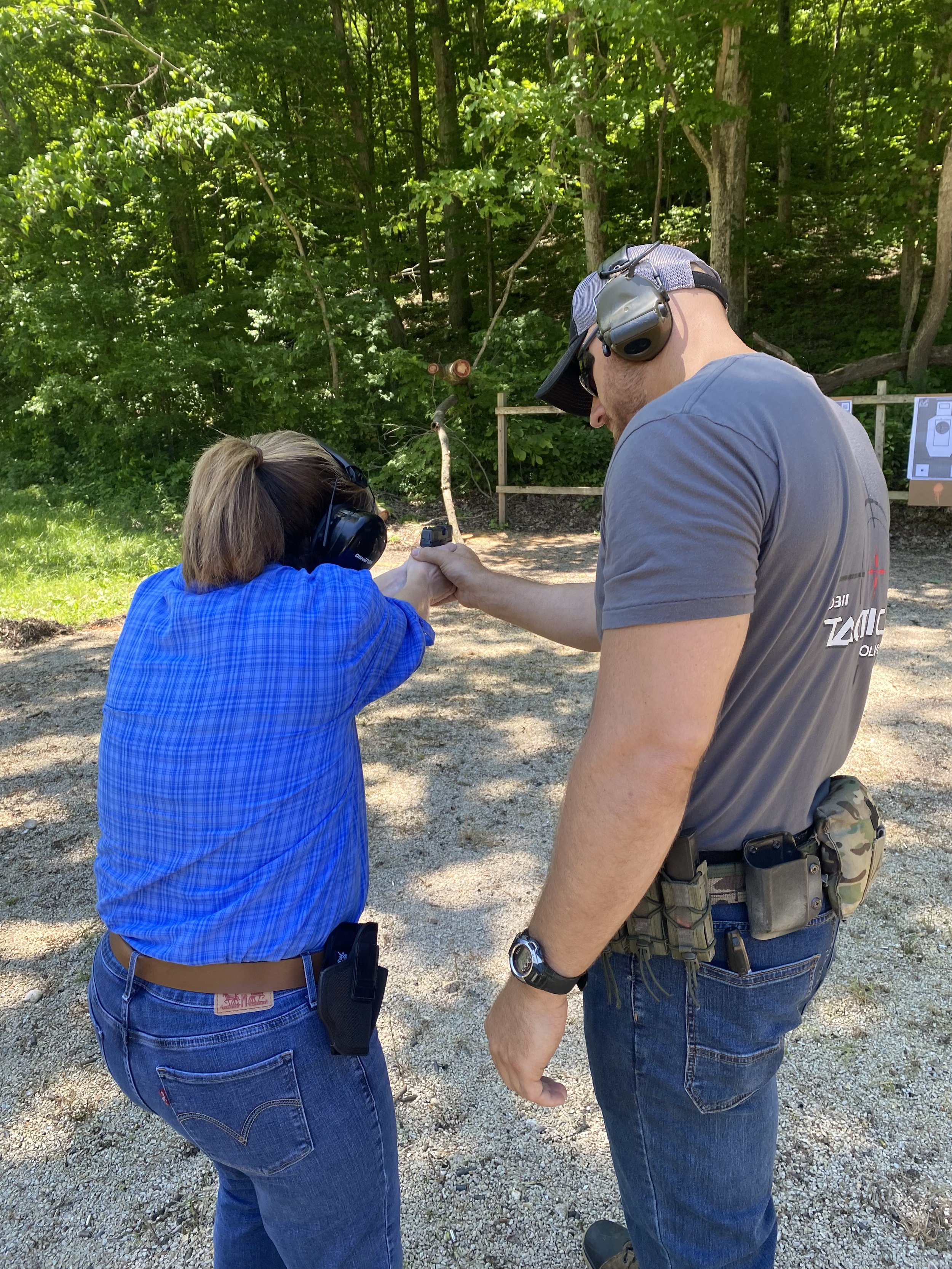 An instructor teaches a woman how to hold and shoot a firearm at a shooting range taking place outdoors in a shaded wooded area.