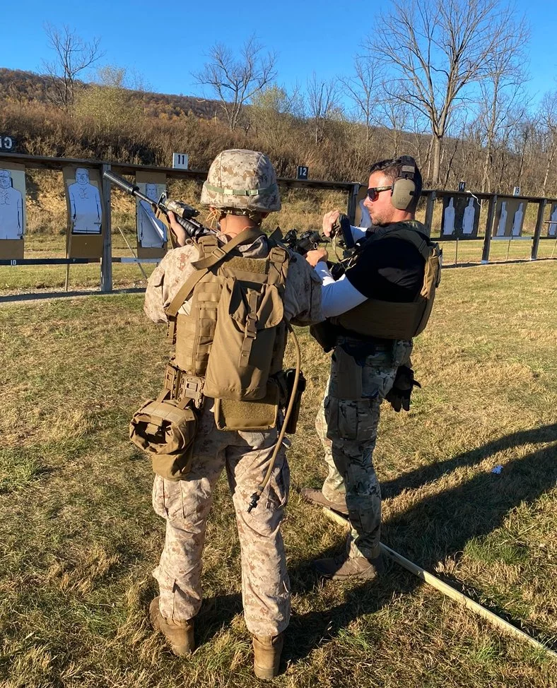 Two men, one in camouflage military gear and the other in tactical clothing, are at an outdoor shooting range during daytime. They are practicing shooting at paper targets placed on stands. The scene features a backdrop of trees and hills with a clea