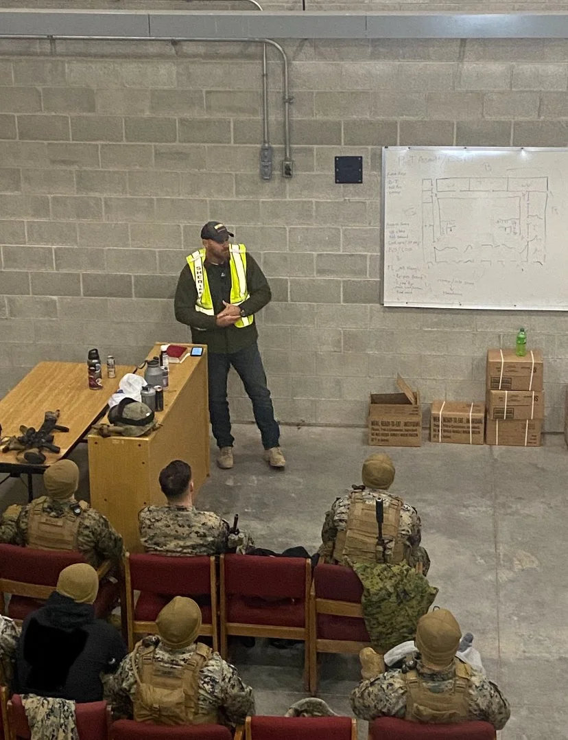 A man in a yellow safety vest and black clothing is standing in front of a group of soldiers in camouflage uniforms, inside a warehouse or training facility, with a whiteboard and boxes visible.