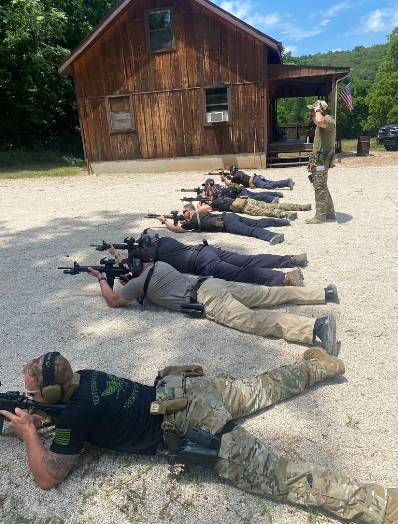 Group of people lying on the ground in a shooting stance with firearms, aiming at a target, outdoors near a wooden building with an American flag.