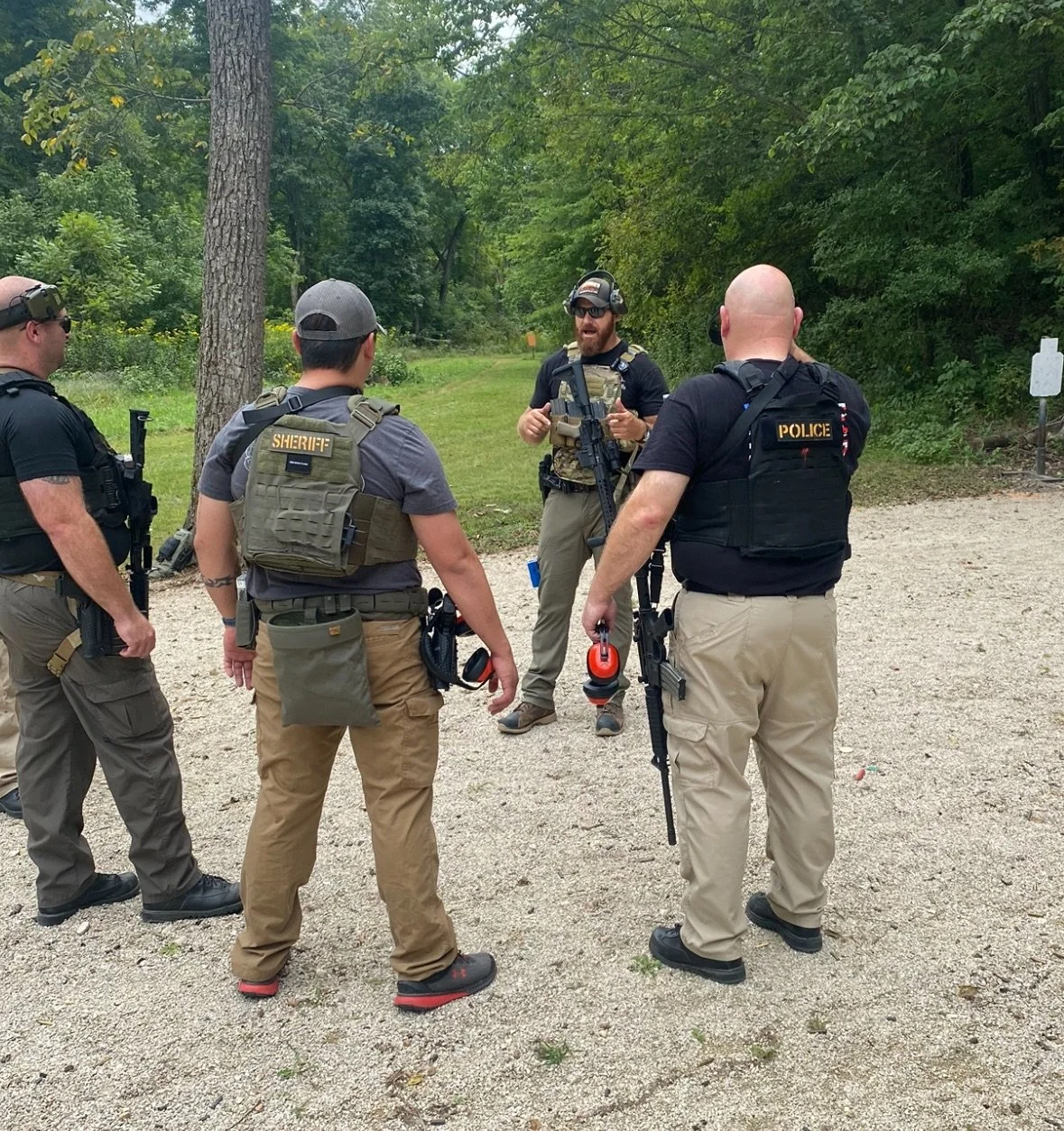 Group of law enforcement officers, including sheriff and police, standing outdoors and having a discussion in a forested area.