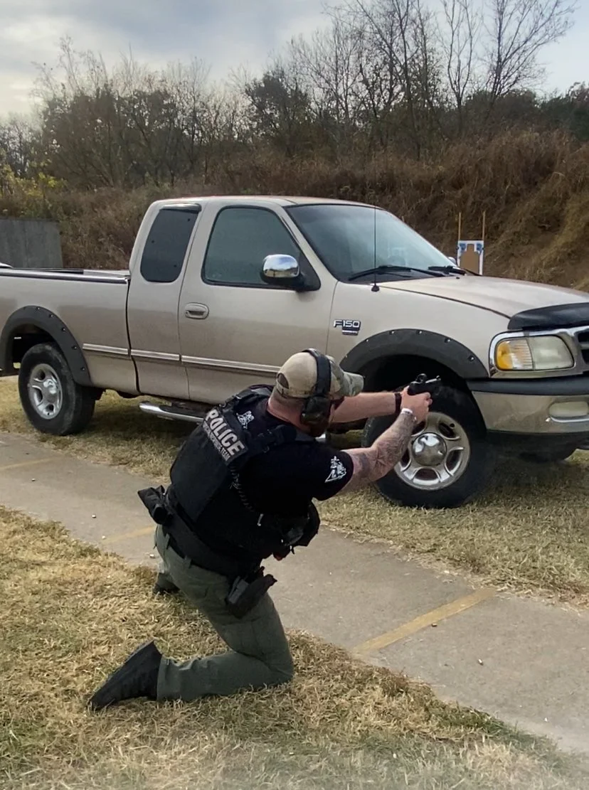 Police officer kneeling on Grass aiming handgun at a pickup truck's tire in an outdoor parking lot