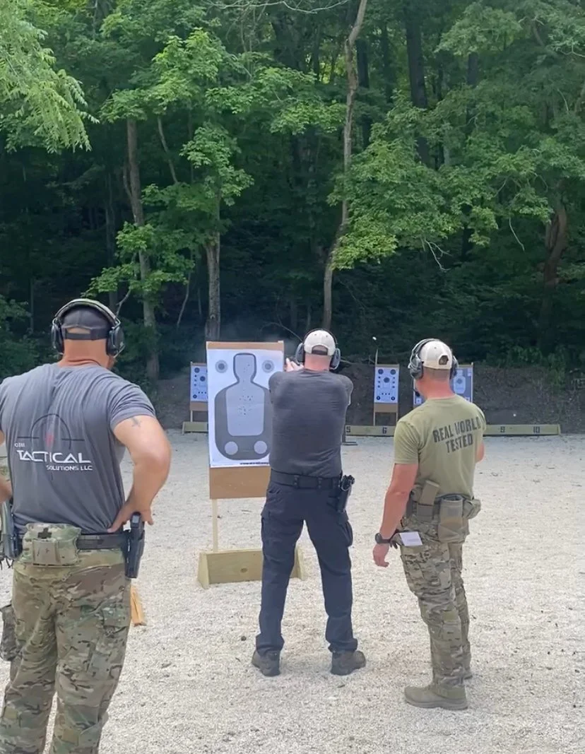 Three men wearing headsets and tactical clothing at an outdoor shooting range, aiming at paper target silhouettes set up in front of a wooded background.