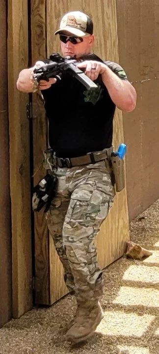 A woman in military-style camouflage pants, a black shirt, and a cap aiming a firearm while standing behind a wooden wall.