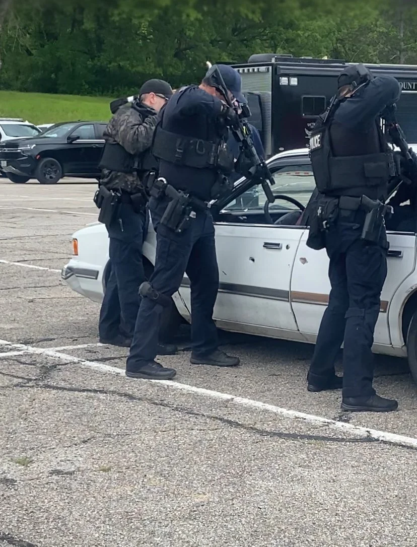 Three police officers in tactical gear inspecting a white car with a roll-up window in a parking lot.