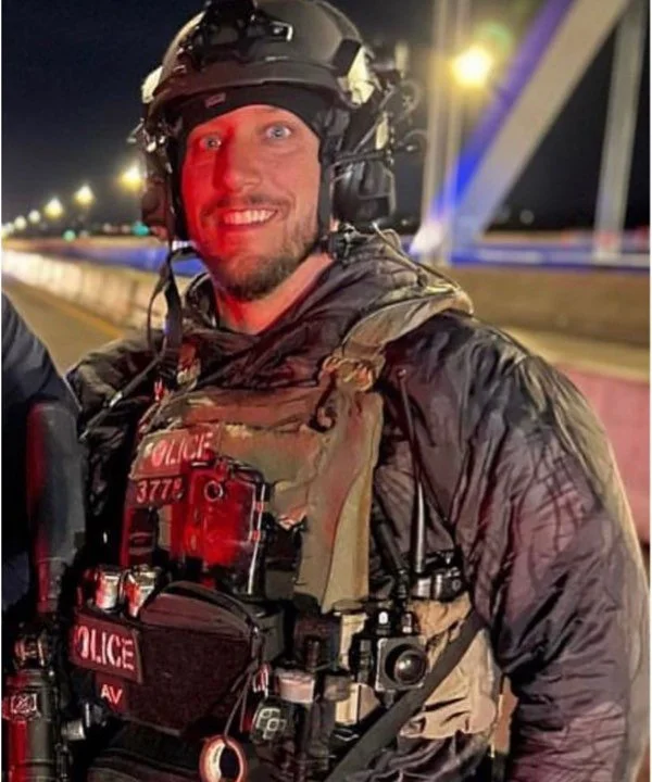 A smiling police officer in tactical gear, including a helmet and a vest, stands outdoors at night with bright city lights and a bridge in the background.