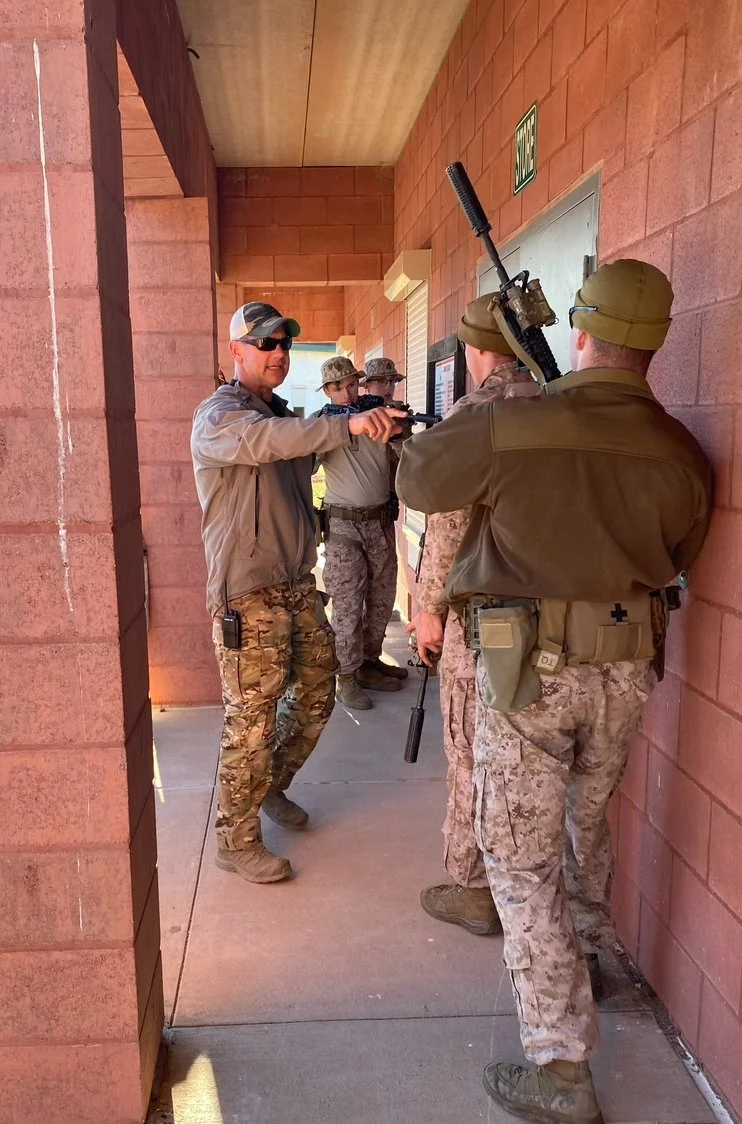 A man in sunglasses and camouflage pants points a handgun at a group of soldiers, some of whom are holding rifles, under a brick building's covered walkway.