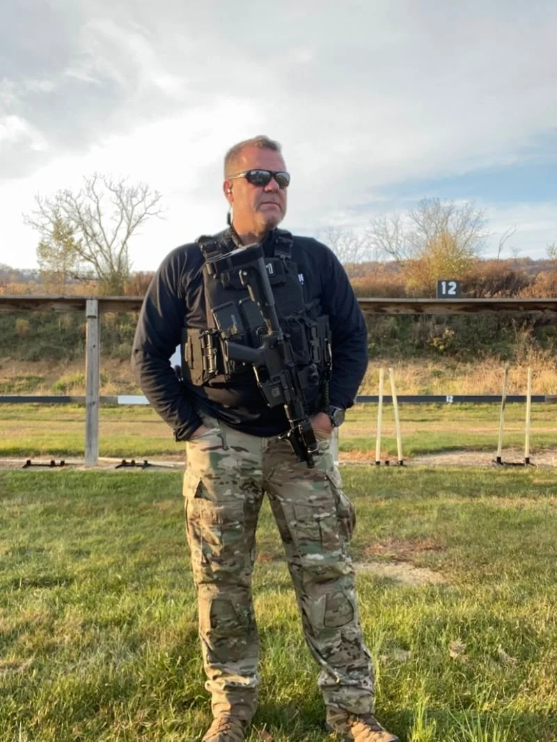 Man in sunglasses, black jacket, camouflage pants, and tactical gear, standing outdoors on grass with a rifle hanging from his chest, under a cloudy sky with some trees and a shooting range backdrop.