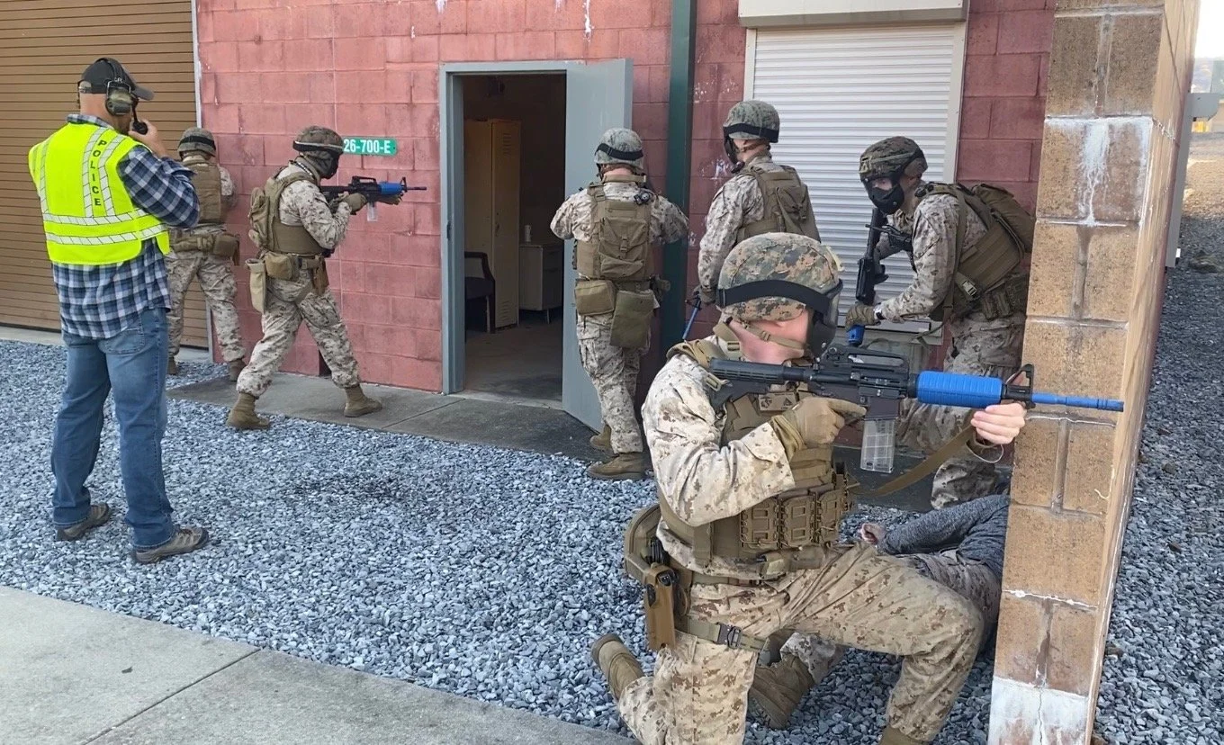 Military personnel training scenario with soldiers in camouflage gear, armed and taking cover in an urban environment.