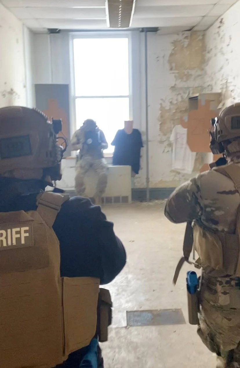 Law enforcement officers in tactical gear inside a room with peeling paint walls and a large window.