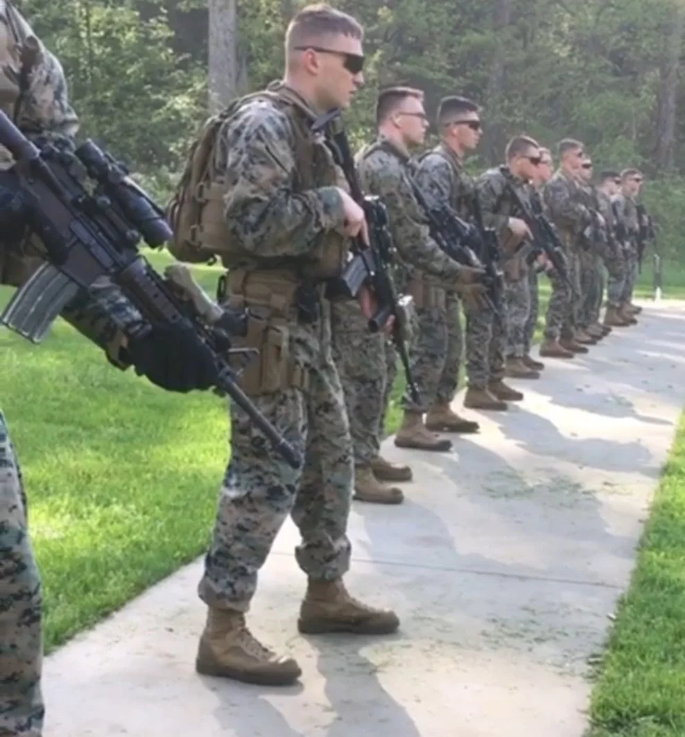 A line of soldiers in camouflage uniforms holding rifles, standing at attention outdoors on a sunny day.