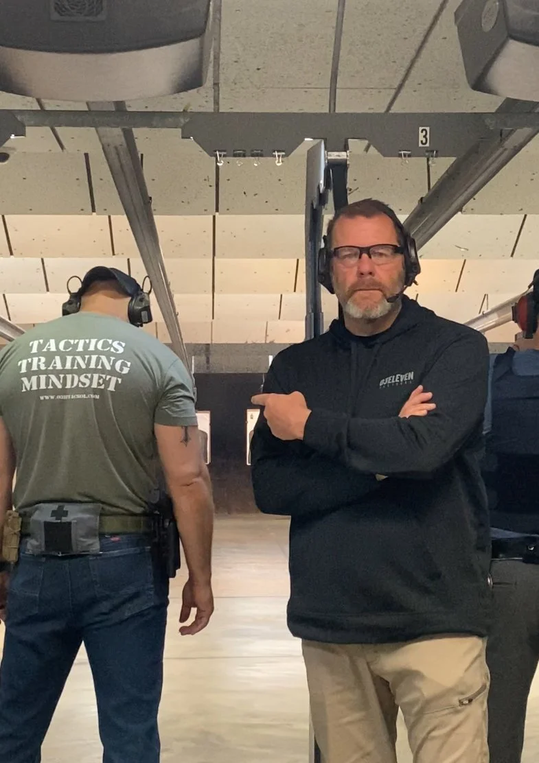 A man with glasses, wearing a black jacket and beige pants, standing with his arms crossed at an indoor shooting range, with other people wearing headphones nearby.