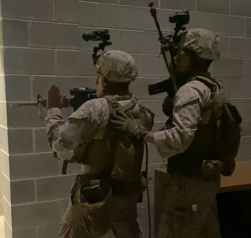 Two soldiers in combat gear aiming rifles around a corner in an indoor setting.