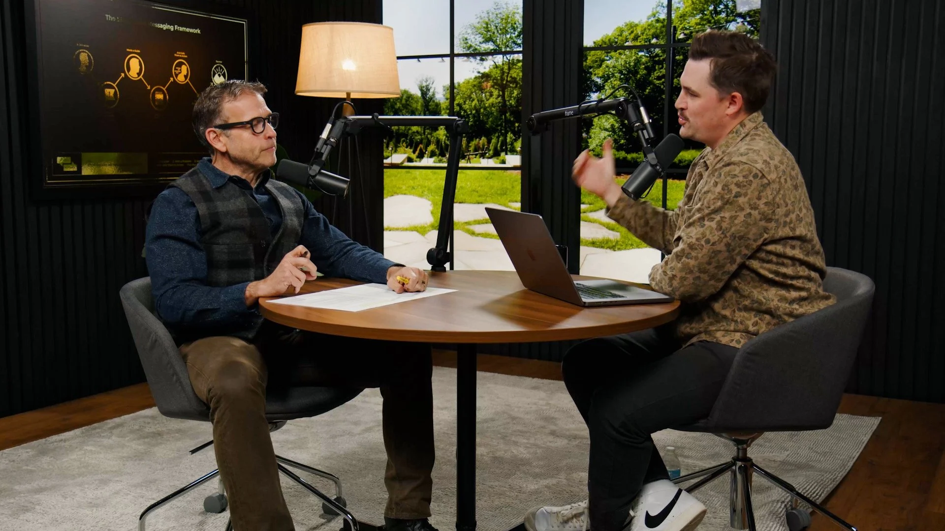 Two men having a discussion during a podcast recording inside a studio with large window and outdoor view, microphones, laptop, and a dark wall with a presentation screen in the new Donald Miller studio.