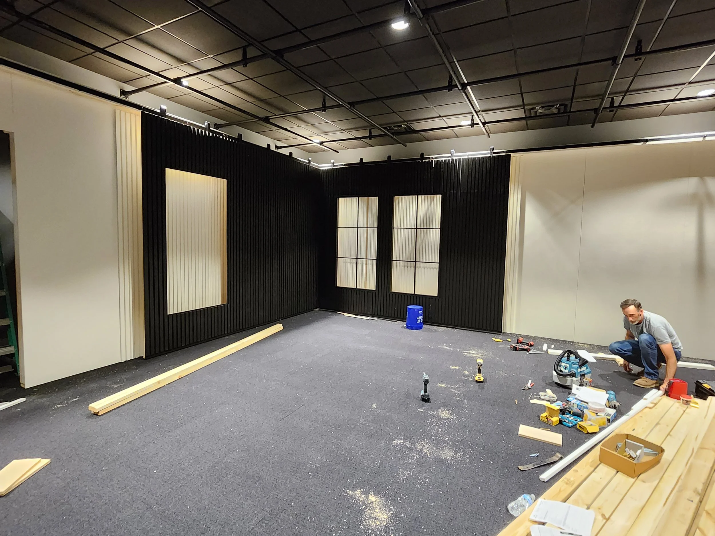 Person working on a construction project inside a room with black and white walls, installing windows or panels, surrounded by tools and materials.