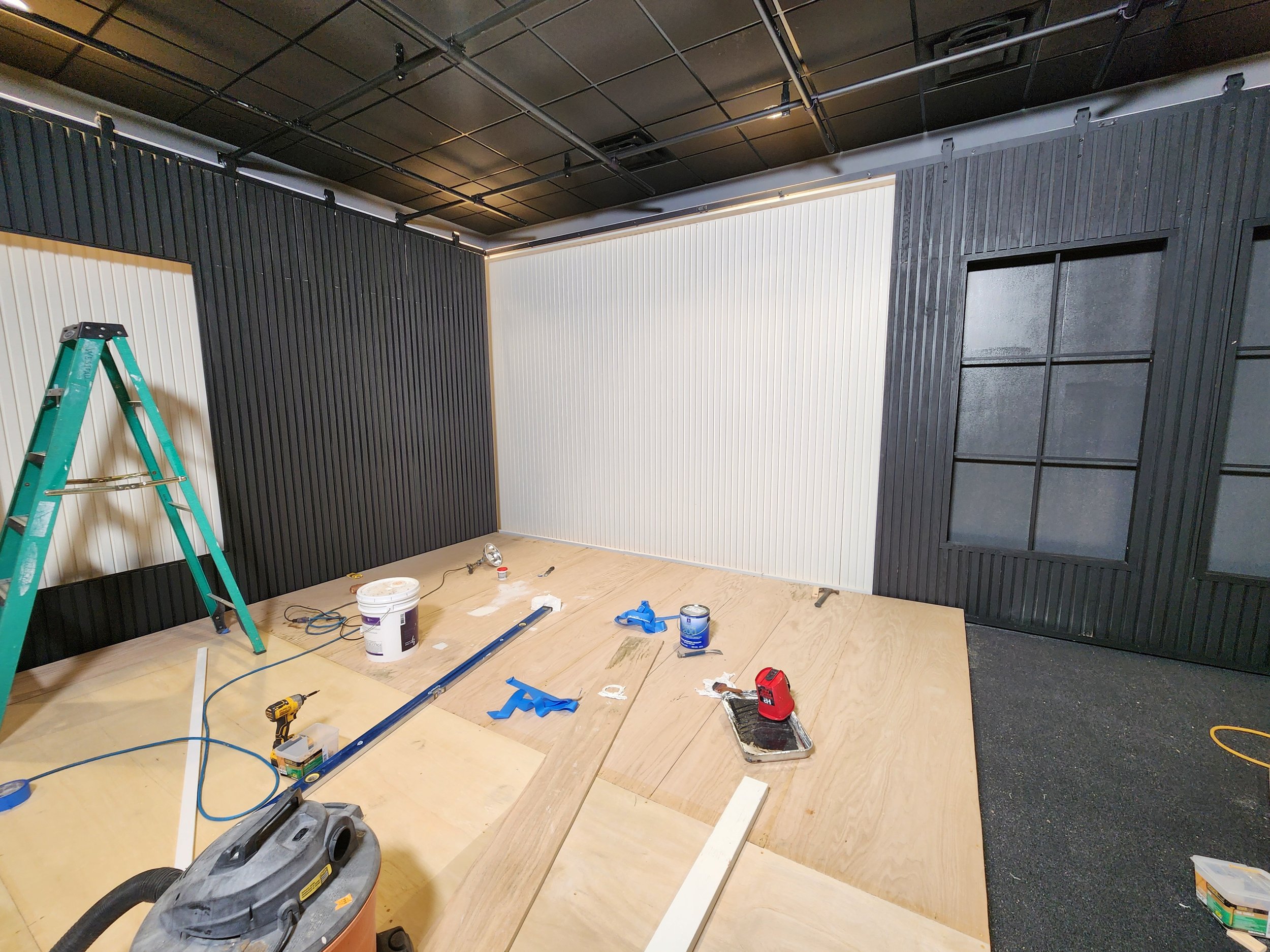 Interior room under construction with black and white paneled walls, a wooden floor, tools, and a green ladder in the new Donald Miller studio.