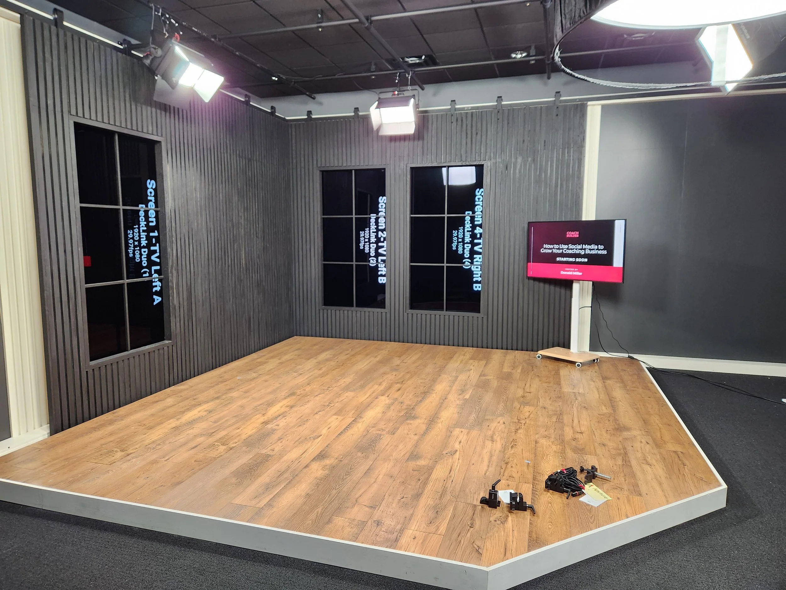 An empty studio stage with a wood floor, black paneled walls, and three television screens on the wall, two displaying setup instructions, with some equipment and cables on the floor and a television monitor on a stand showing a presentation slide.