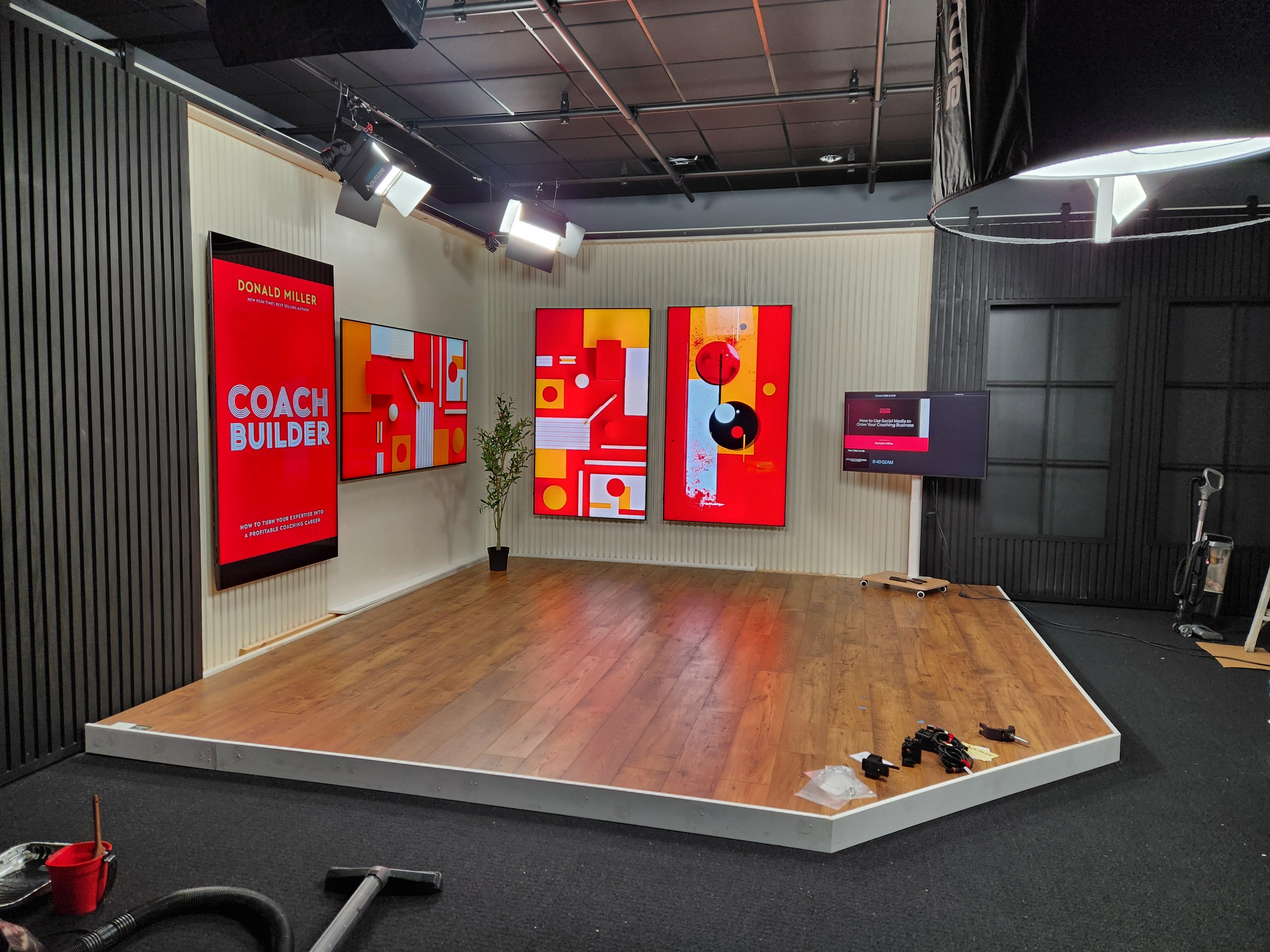 A studio with a small wooden stage and a wall decorated with digital screens displaying a presentation titled 'Coach Builder' with a red, yellow, and white abstract background. To the right is a mounted TV monitor showing a slide in the new studio.