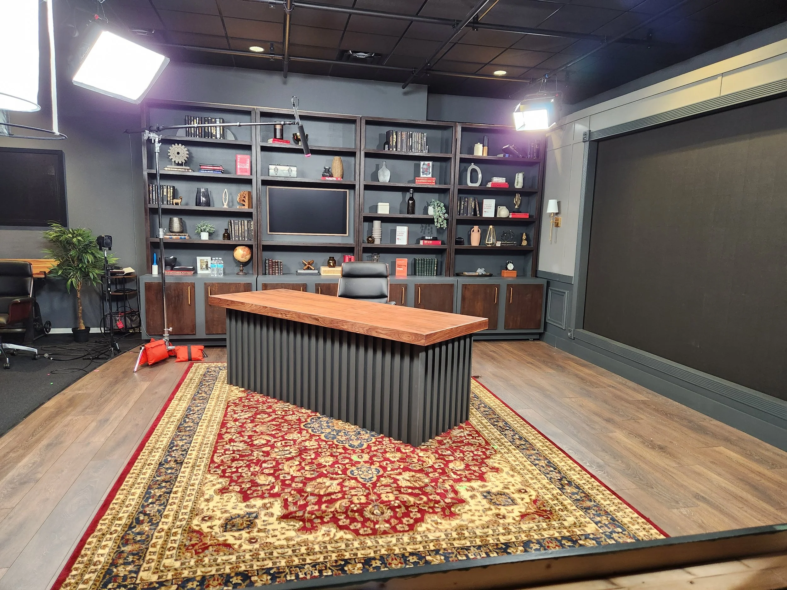 An empty television or interview studio with a wooden desk on a red and cream patterned rug, a large gray bookshelf with decorations, plants, and a large black backdrop on the wall.