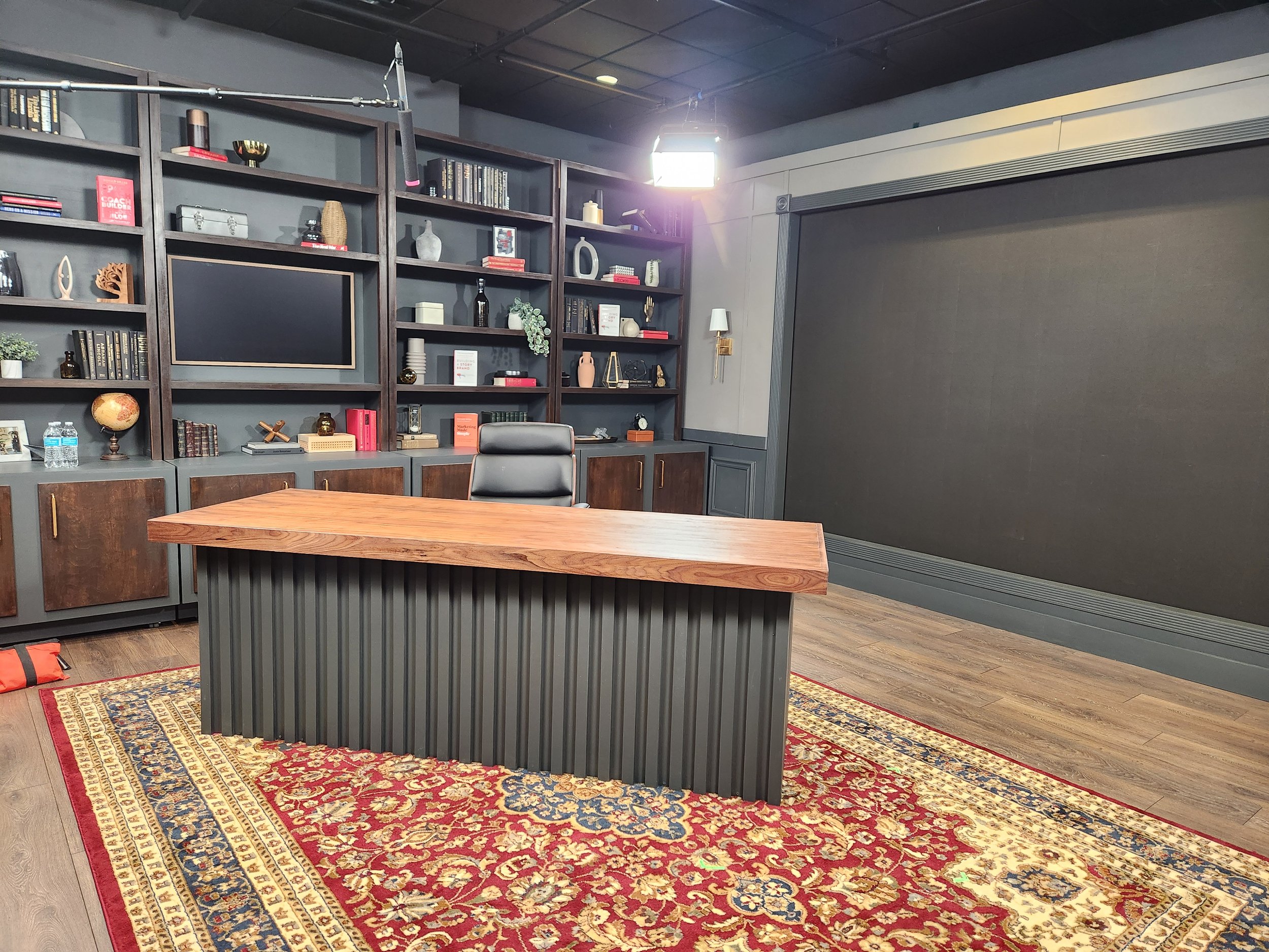 An office with a wooden desk, black leather chair, and a decorative area rug. Green bookshelves with books and ornaments are in the background, along with a large black screen or wall in the NEW Donald Miller studio.