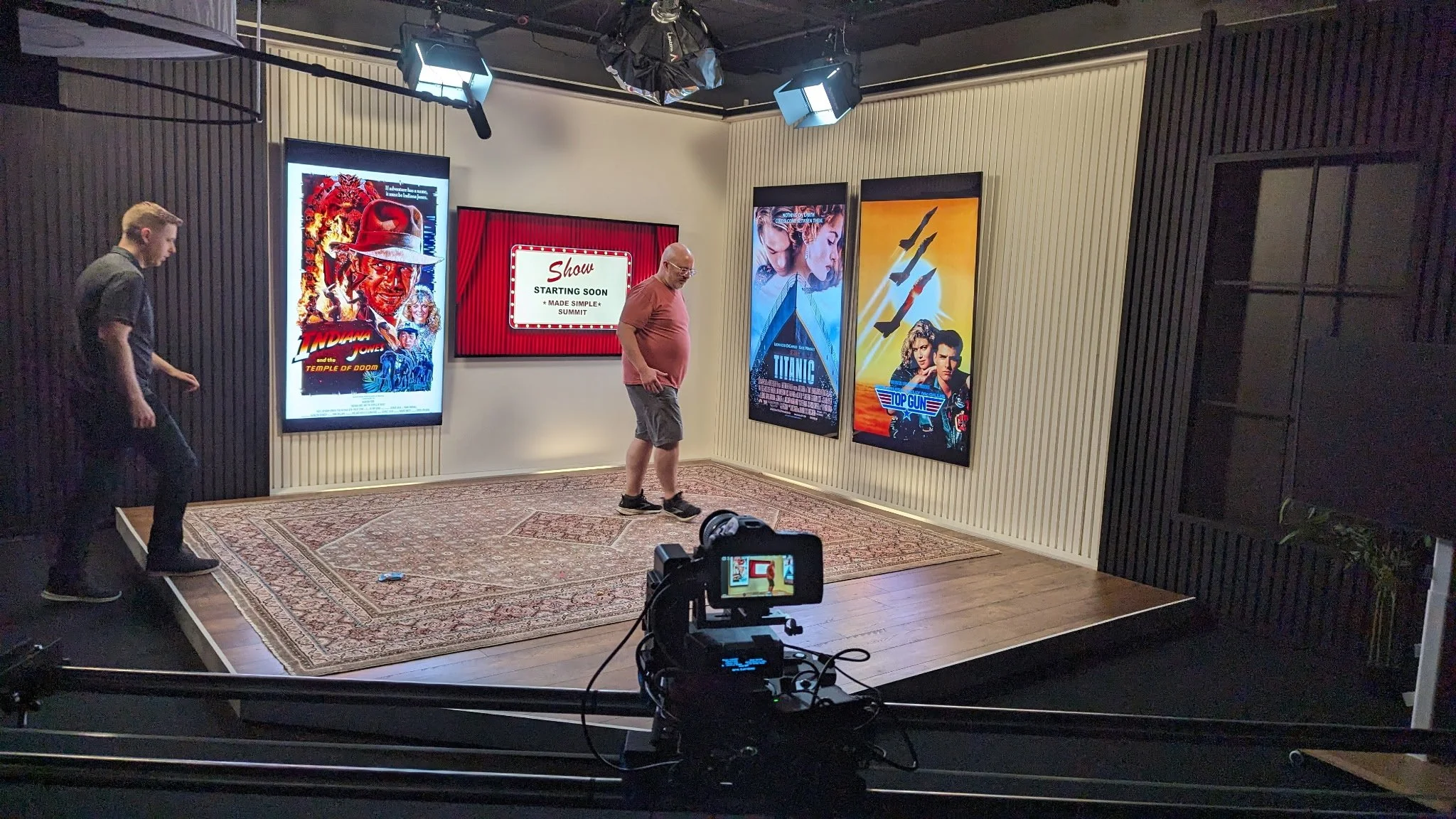 Television studio with a small stage, posters of movies like Indiana Jones, Titanic, Top Gun, and a screen that says 'Show Starting Soon.' Two men are setting up on stage, one adjusting his position. Professional lighting equipment hangs from the cei