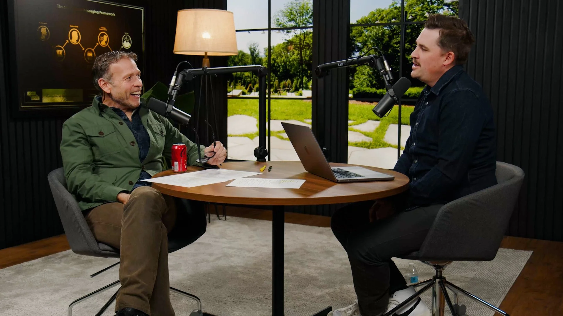 Two men having a conversation in a podcast studio, with microphones, a laptop, papers, and a red soda can on the table, and a large window showing a green outdoor scene, in the new Donald Miller studio.