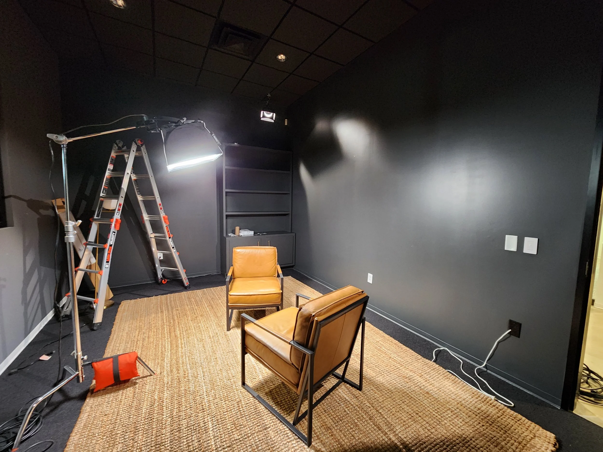 Setup for a video or photo shoot with two tan chairs on a woven area rug, ladder and lighting equipment in a dark room with black walls in the new Donald Miller studio.