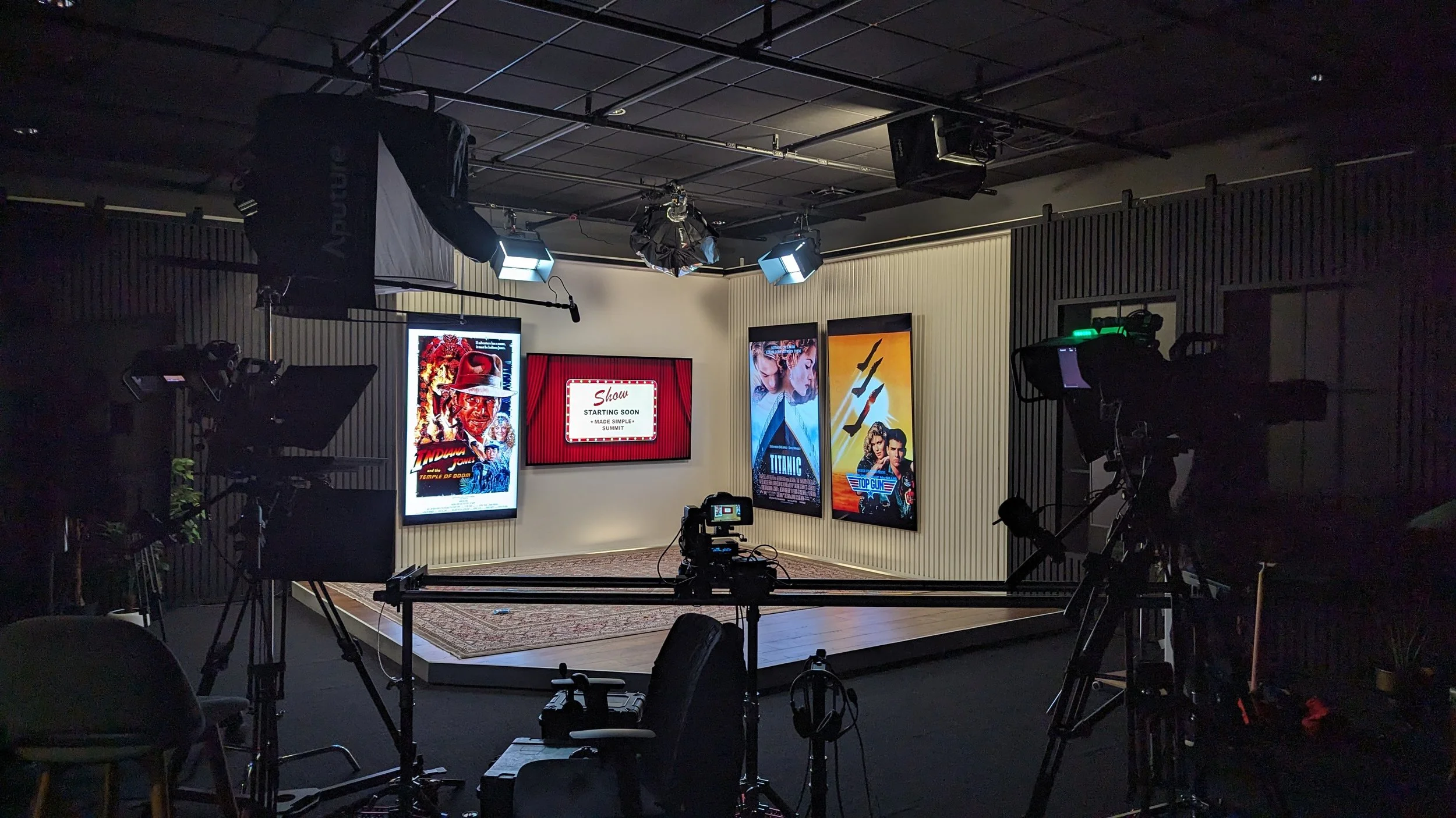 Television studio with cameras, lighting, and a small stage displaying movie posters, including Indiana Jones, Titanic, and Top Gun, with a screen that says "Show starting soon".