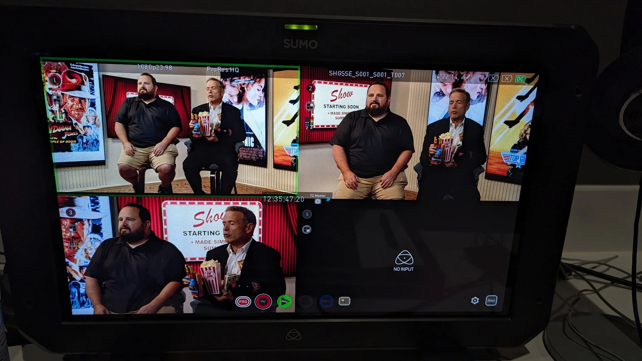 A television screen shows a behind-the-scenes shot of a video production with two men seated in front of a red curtain and television posters, one holding a bucket of popcorn, and a third man standing in front of the curtain, in a studio setting.