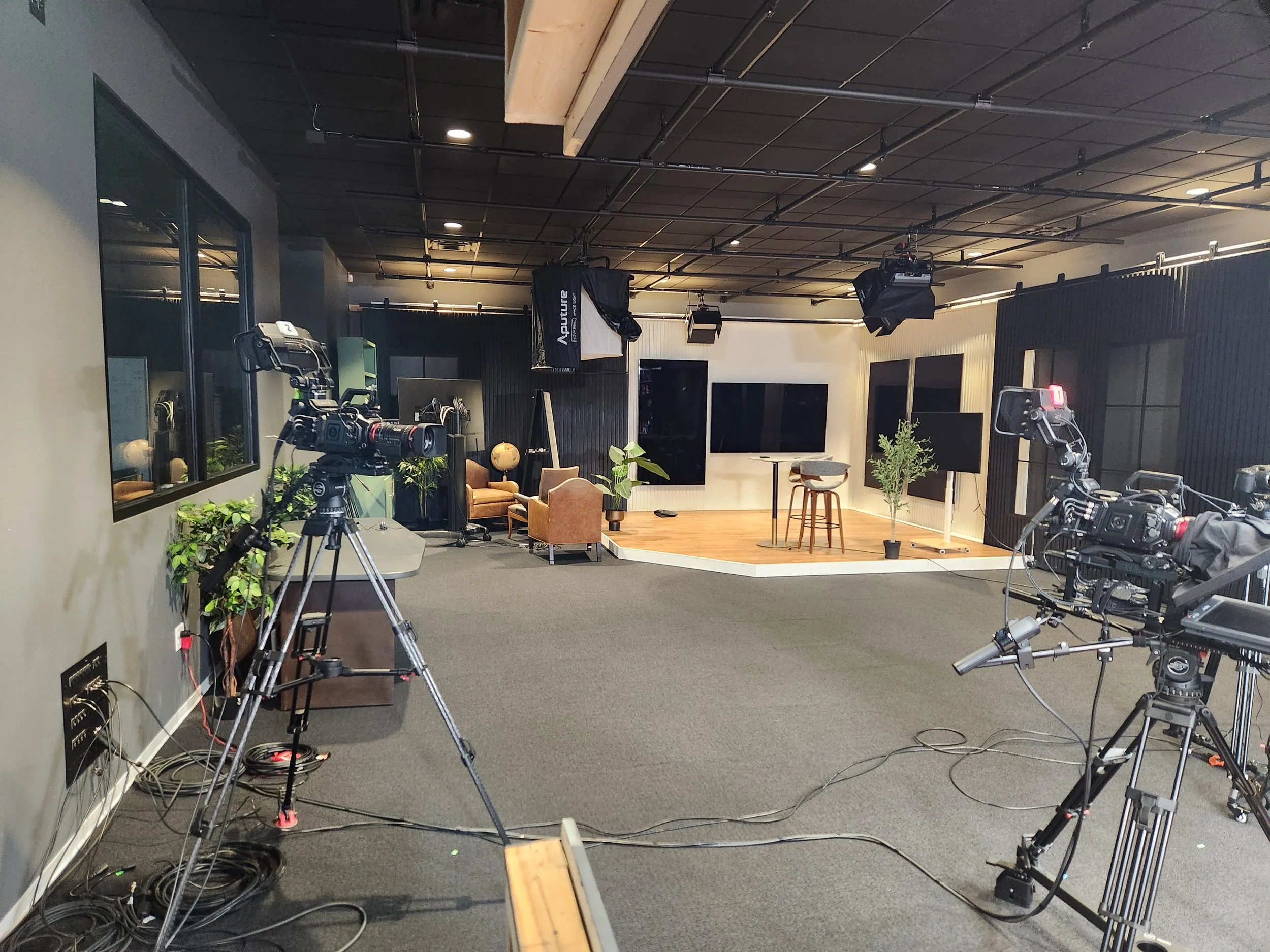A television studio with cameras, lighting equipment, and a small stage featuring chairs, plants, and screens.