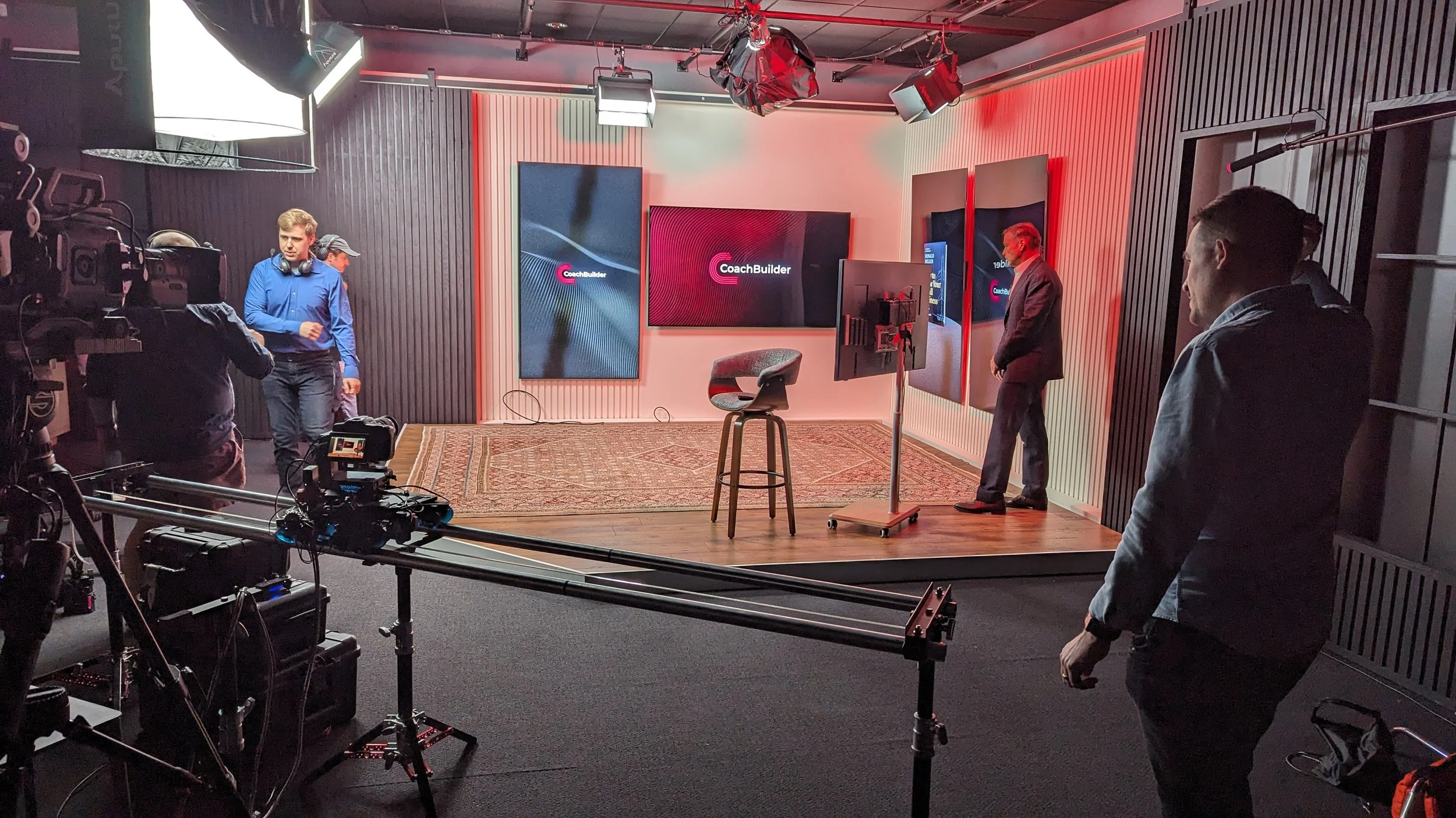 Behind-the-scenes view of a video studio setup with three men, professional camera equipment, four screens displaying the CoachBuilder logo, and a stool in front of the screens in the new Donald Miller studio.