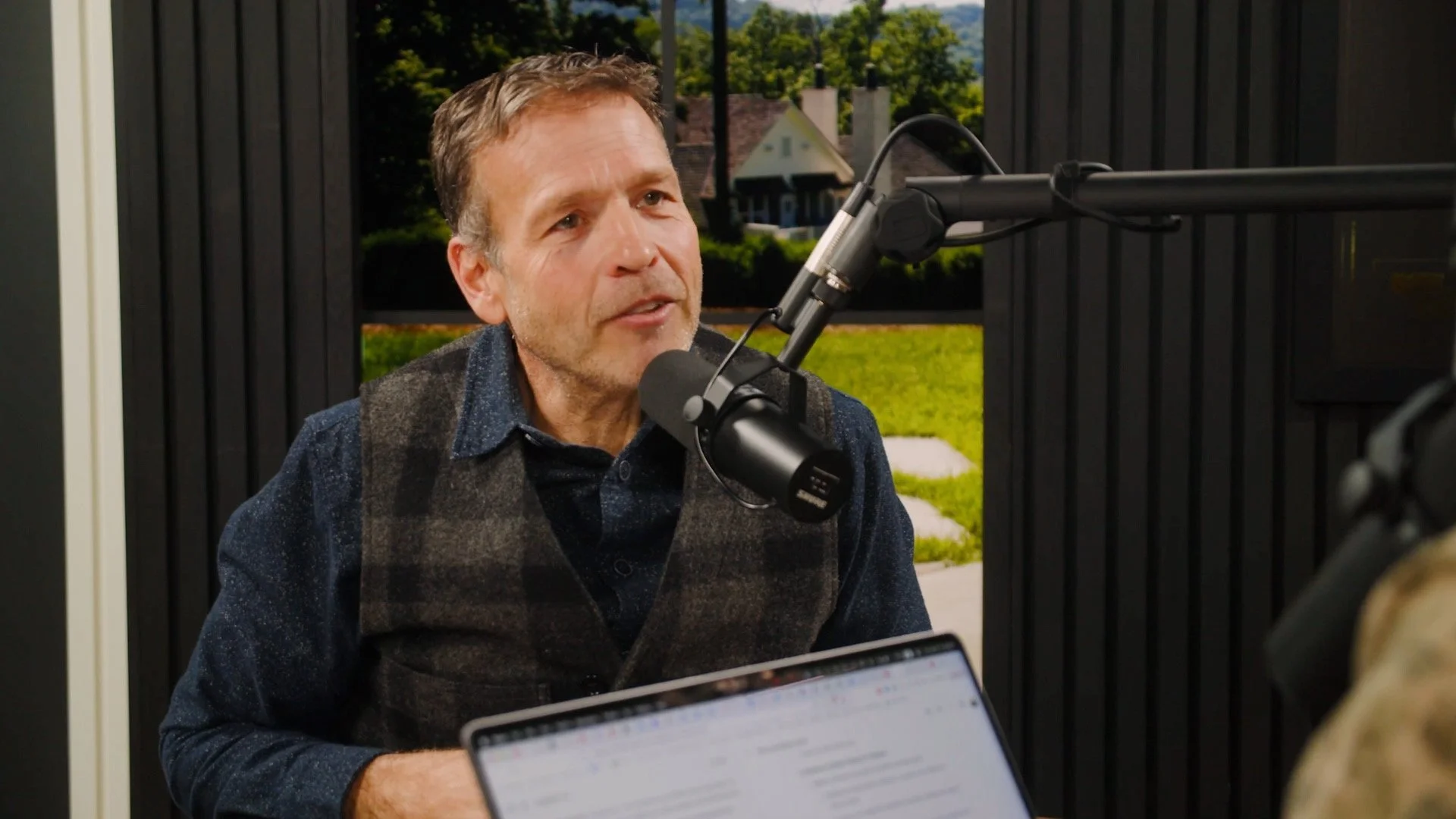 A man with brown hair and a beard, wearing a dark patterned shirt and vest, speaking into a microphone during a recording session with a scenic outdoor background visible through the window in the new Donald Miller studio.