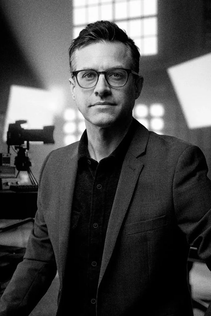 Black and white photo of a man with glasses sitting in a photography studio with professional lighting and equipment.