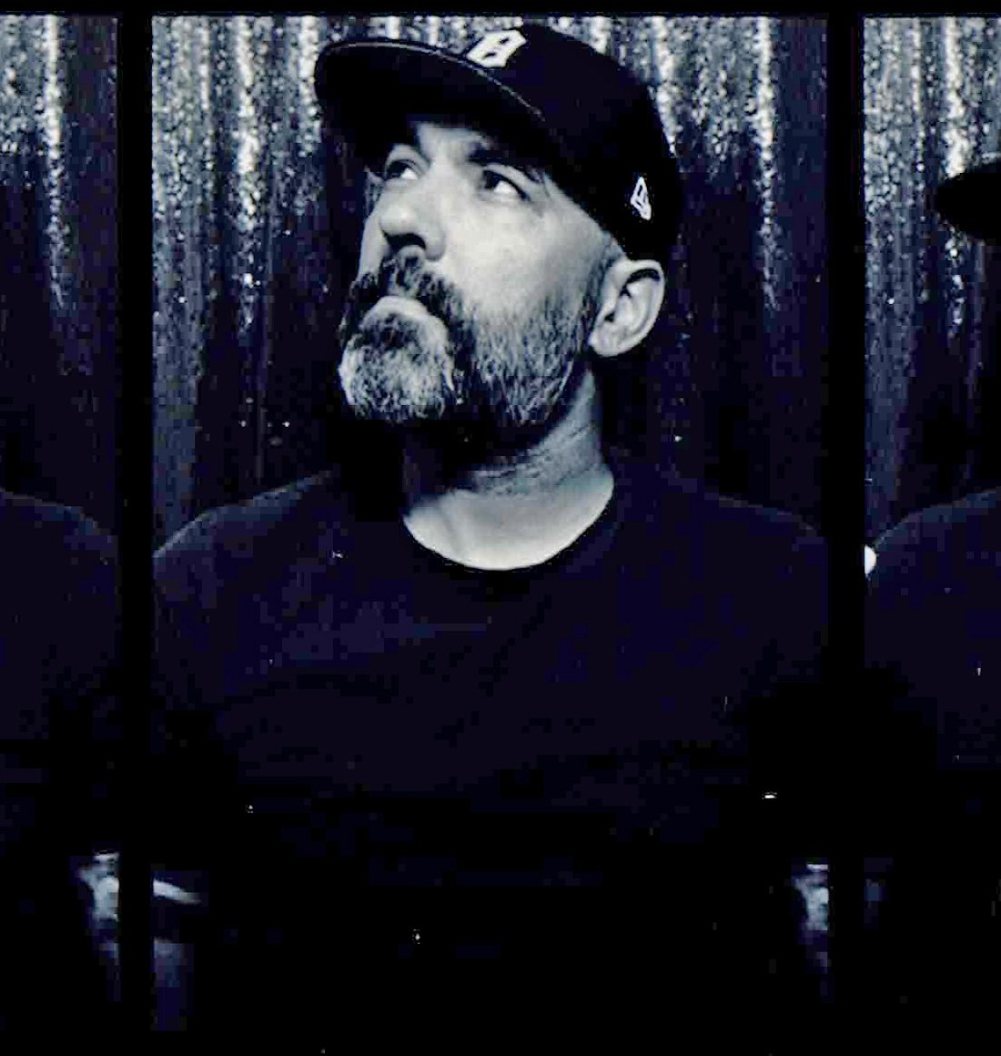 Black and white portrait of a man with a beard wearing a baseball cap, looking upwards, behind metal bars.