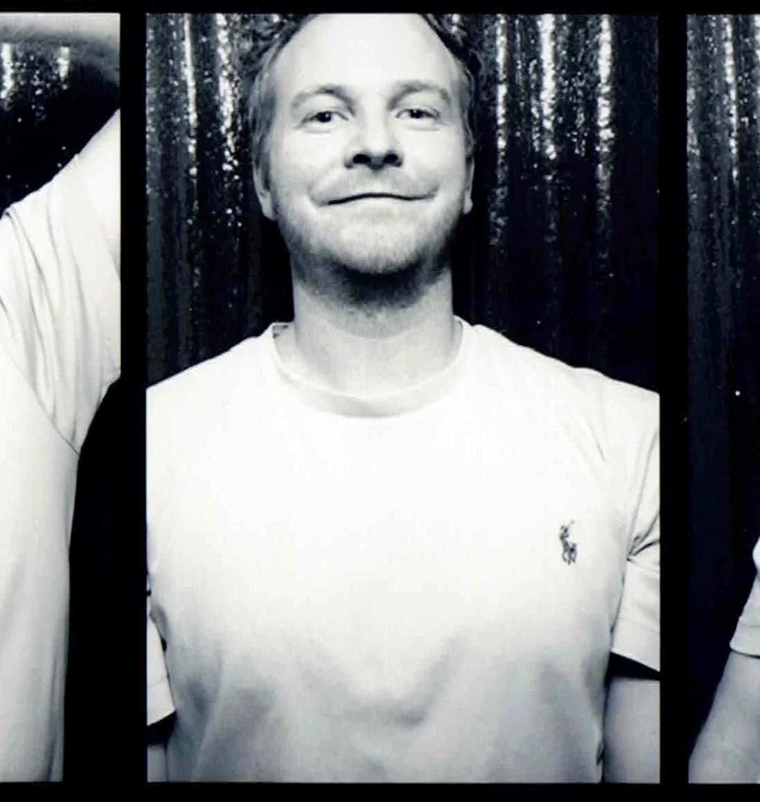 Black and white photo of a smiling man wearing a white T-shirt with a small logo, standing in front of a shiny, textured curtain.