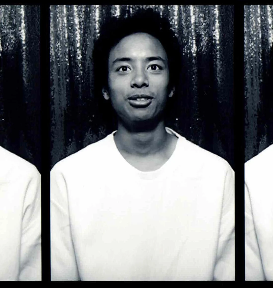 Black and white photo of a young man with dark, curly hair, wearing a white shirt, standing in front of a glittery, textured curtain background.