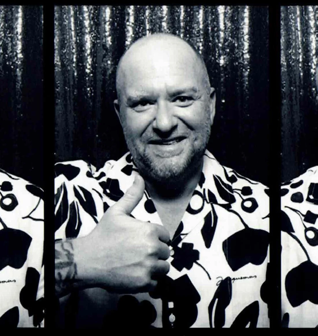 A man with a shaved head and beard giving a thumbs up, wearing a patterned shirt, standing behind black bars with a sparkly curtain in the background.