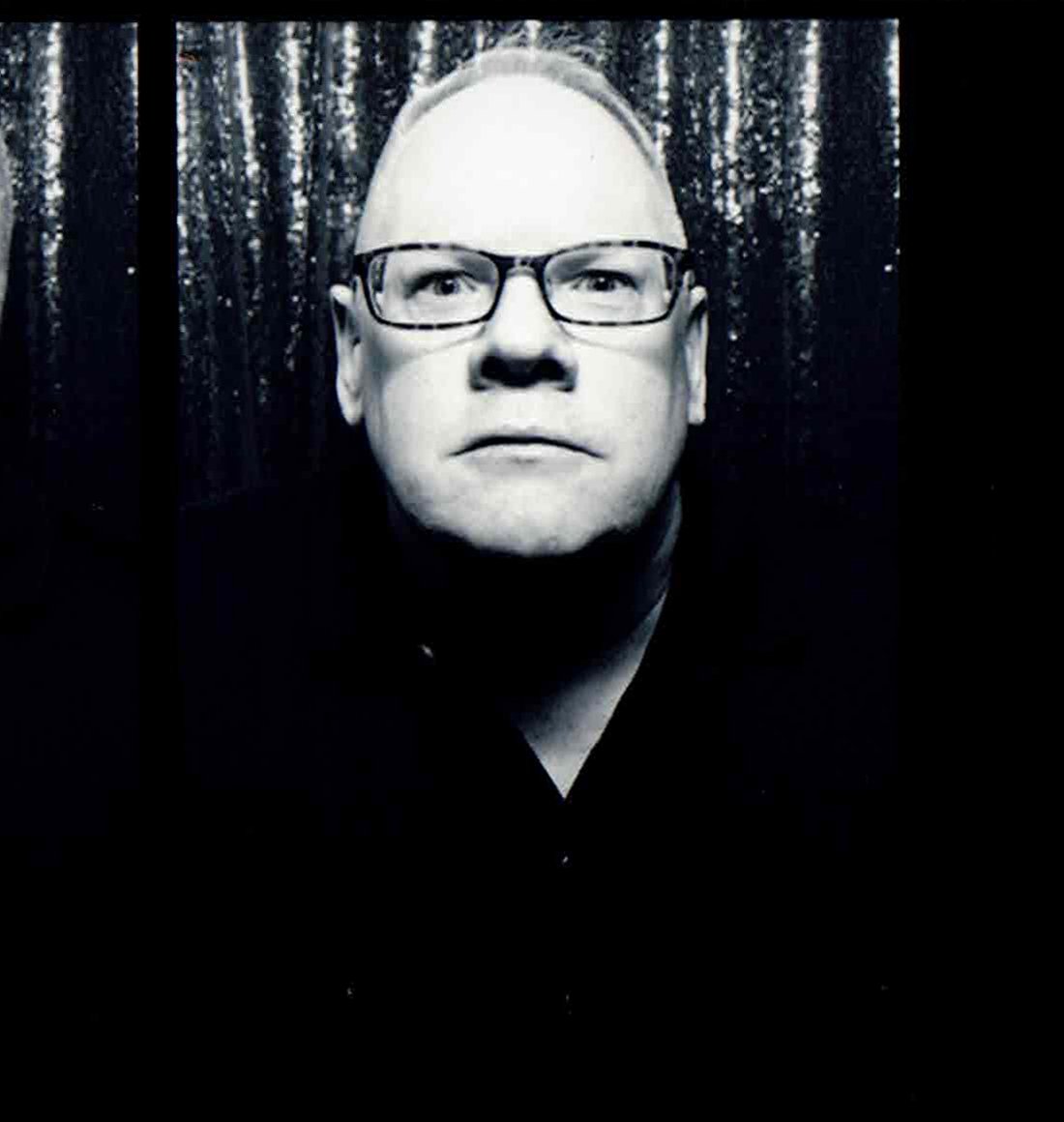 Black and white portrait of a man wearing glasses, with a serious expression, in front of a dark, textured background.