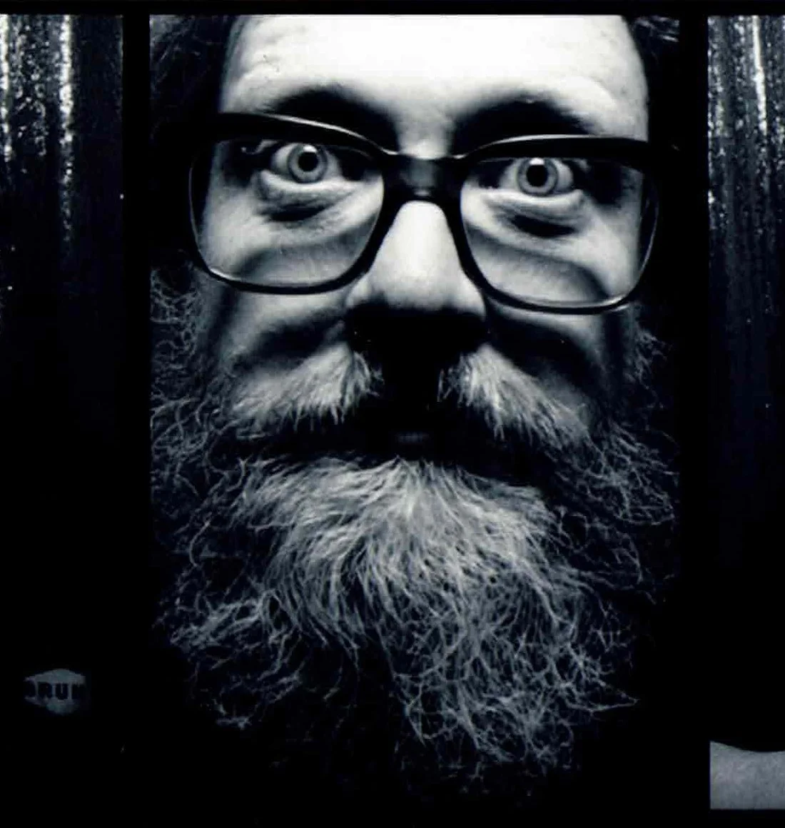 Close-up black and white photo of a man's face with glasses, gray beard, and mustache, looking directly at the camera.