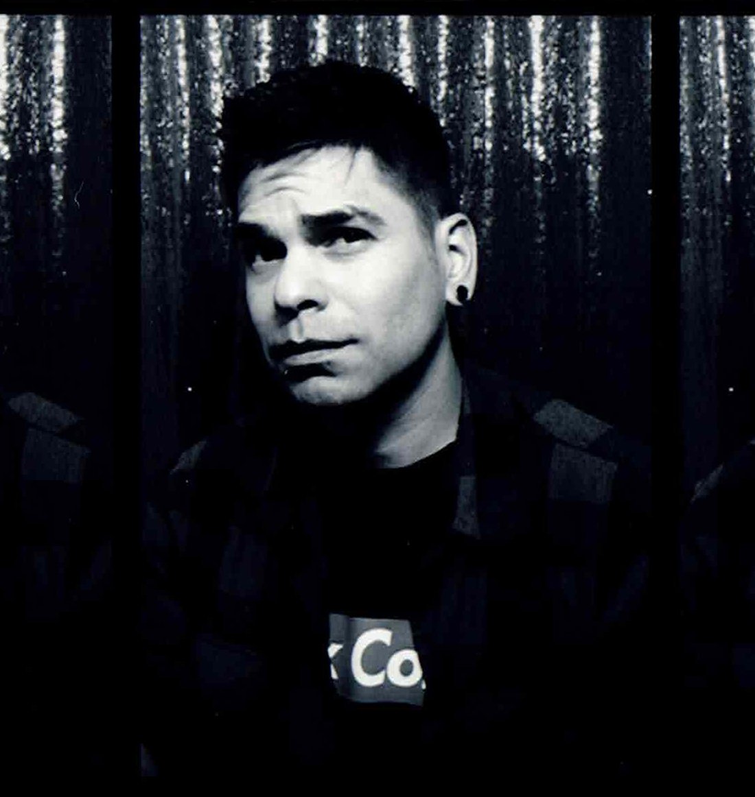 Black and white photo of a young man with short dark hair and earrings, looking to the side, standing in front of a forest background.